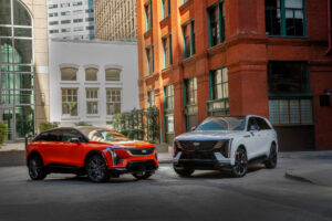 Front 3/4 view of the 2025 Cadillac ESCALADE IQ in Summit White and 2025 Cadillac OPTIQ in Monarch Orange parked on a city street.
