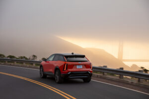 Rear 3/4 view of the 2025 Cadillac OPTIQ in Monarch Orange driving on a mountain road with a scenic view in the background.